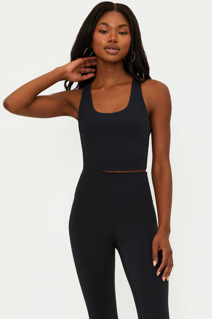 A female model wearing a black matte longline crop top with a scoop neckline and a racerback design.