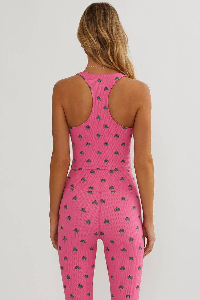 A woman modeling a matching two-piece activewear set consisting of a pink sports bra and high-waisted leggings, both featuring a repeating dark pink strawberry print, against a plain light gray studio background.