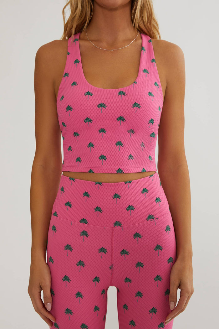 A woman wearing a matching pink activewear set with a small dark green palm tree print, featuring a scoop-neck sports bra and high-waisted leggings, shown against a plain light gray background.