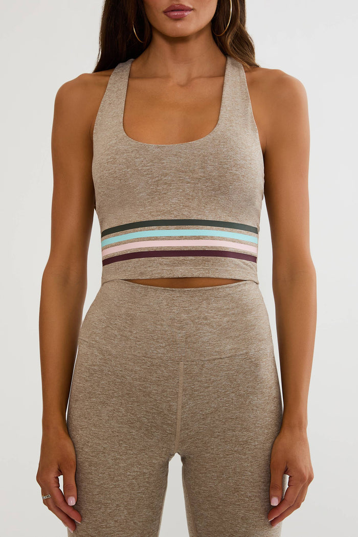 A woman wearing a heather taupe athletic set consisting of a scoop-neck crop top with a multicolor striped waistband and matching high-waisted leggings, shown against a plain light gray background.