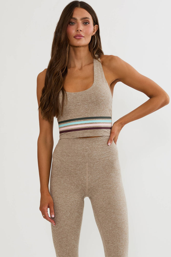 A woman modeling a matching beige activewear set consisting of a halter-neck sports bra and high-waisted leggings, featuring a horizontal striped waistband in black, white, light blue, and burgundy.